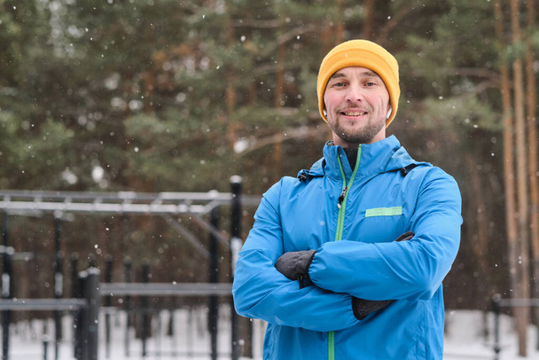 Portrét usmívajícího se mladého sportovce v teplém saku, stojícího se zkříženýma rukama pod padajícím sněhem na cvičišti - Fotografie, Obrázek