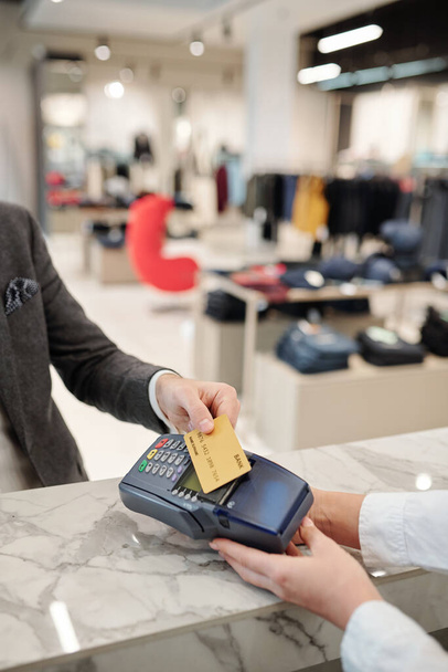 Close-up of unrecognizable businessman standing at counter in clothing store and putting credit card to payment terminal - Photo, Image