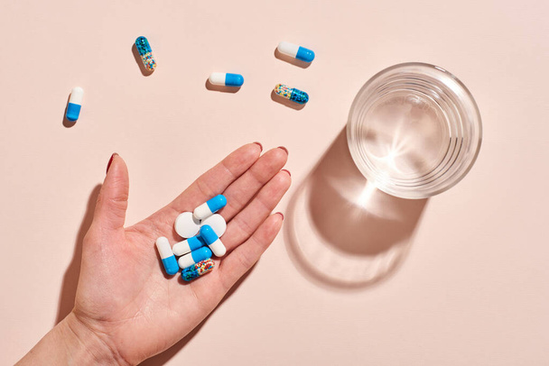 Various medicaments in women's hand with glass of water on pale pink background, horizontal flat lay shot - Foto, afbeelding