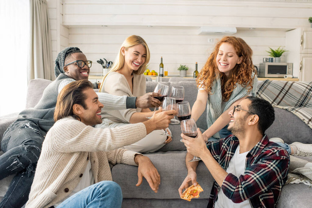 Tiro horizontal de alegres hombres y mujeres jóvenes pasando el fin de semana juntos en acogedor apartamento tintineo vasos con vino
 - Foto, Imagen