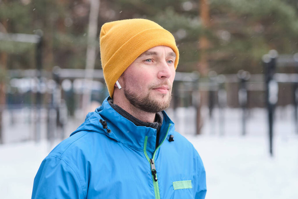 Pensive motivated young sportsman with stubble listening to music in ear buds while standing outdoors in winter - Foto, Bild