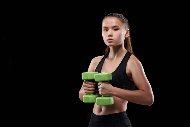 Ajuste jovem mulher em sportswear segurando halteres pelo peito enquanto faz exercício contra fundo preto
 - Foto, Imagem