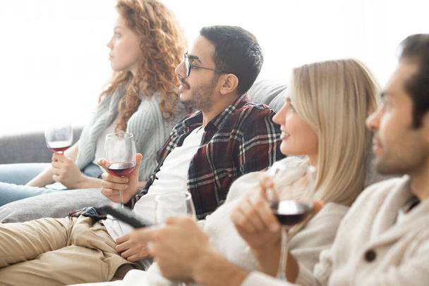 Group of young multi-ethnic friends with wineglasses sitting on sofa and watching tv together - Фото, зображення