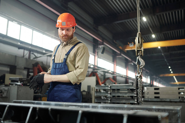 Ingeniero barbudo en ropa de trabajo y hardhat parado por el detalle enorme del hierro mientras que controla el proceso de trabajo en fábrica
 - Foto, Imagen