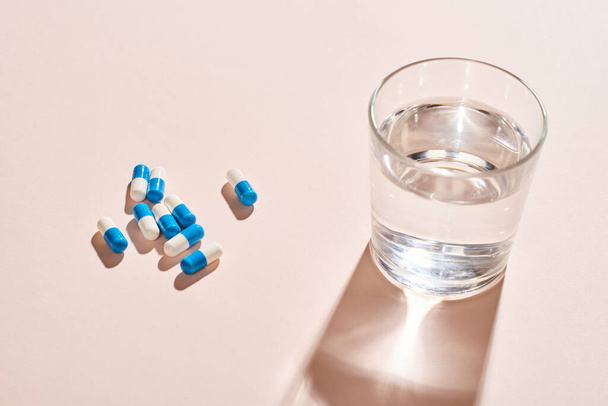 White And blue capsule pills and glass of water on pale pink table surface, horizontal shot - Foto, Imagen