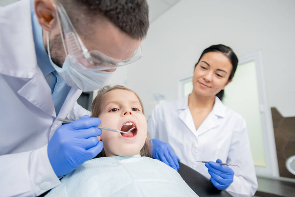 Junge Zahnärztin mit Zahnspiegel während der oralen Untersuchung eines kleinen Mädchens, das vor dem Hintergrund eines Assistenten den Mund offen hält - Foto, Bild