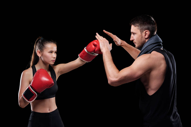 Młoda, sprawna kobieta w ubraniu aktywnym i rękawicach bokserskich uderzająca trenera podczas walki z czarnym tłem - Zdjęcie, obraz