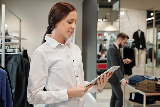 Lächelnder junger Angestellter eines Bekleidungsgeschäfts sucht mit Tablet nach Verkäufercode, während Geschäftsmann im Hintergrund Kleidung auswählt - Foto, Bild