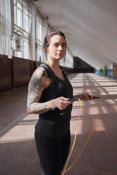 Jovem atleta feminina tatuada em sportswear preto de pé com pular corda no corredor e olhando para a distância pensativa
 - Foto, Imagem