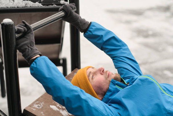 Σοβαρός νεαρός άνδρας ξαπλωμένος σε πάγκο άσκησης εξωτερική και κρατώντας οριζόντια γραμμή, ενώ κάνει crunches στην προπόνηση προπόνηση το χειμώνα - Φωτογραφία, εικόνα
