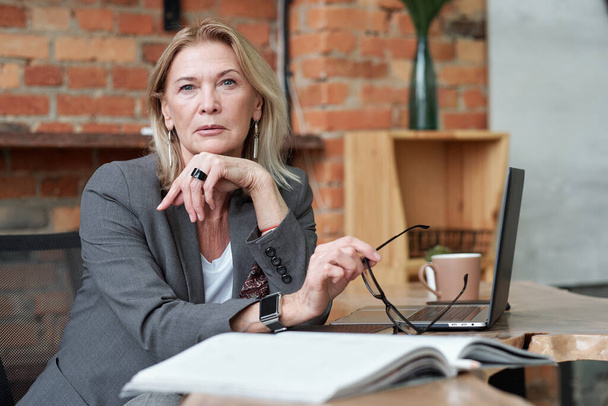 Akıllı saati olan ciddi bir kadın CEO 'nun portresi masada oturuyor ve elinde gözlük tutuyordu. - Fotoğraf, Görsel