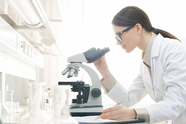 Horizontal side view shot of young female medical scientist examining specimen using microscope in modern laboratory - Foto, afbeelding