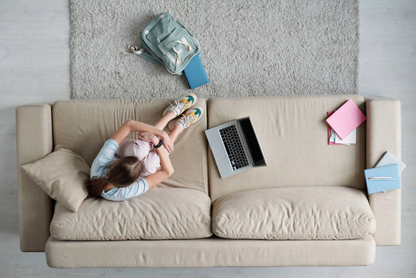 Вид зверху депресивна школярка сидить на дивані з ноутбуком і книгами у вітальні і обличчям при плачу
 - Фото, зображення