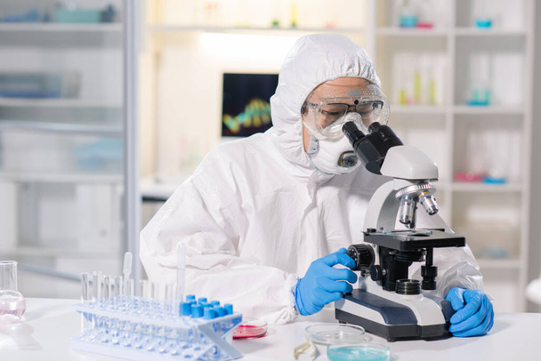 Ασιάτης ιατρικός επιστήμονας στον αναπνευστήρα και γυαλιά ασφαλείας χρησιμοποιώντας μικροσκόπιο για την έρευνα μολυσμένου δείγματος αίματος στο εργαστήριο - Φωτογραφία, εικόνα