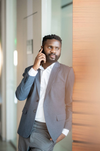 Conteúdo confiante jovem empresário negro de jaqueta em pé na parede e falando por telefone celular
 - Foto, Imagem