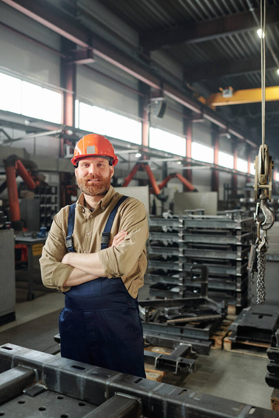 Νεαρός επιτυχημένος διασταυρωμένος μηχανικός που σε κοιτάζει μέσα στο βιομηχανικό εργοστάσιο πάνω σε στοίβες από τεράστιες λεπτομέρειες - Φωτογραφία, εικόνα