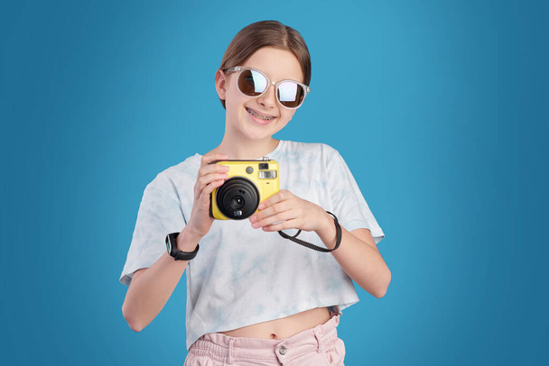 Portret van positief tienermeisje in zonnebril fotograferen op instant camera tegen blauwe achtergrond - Foto, afbeelding