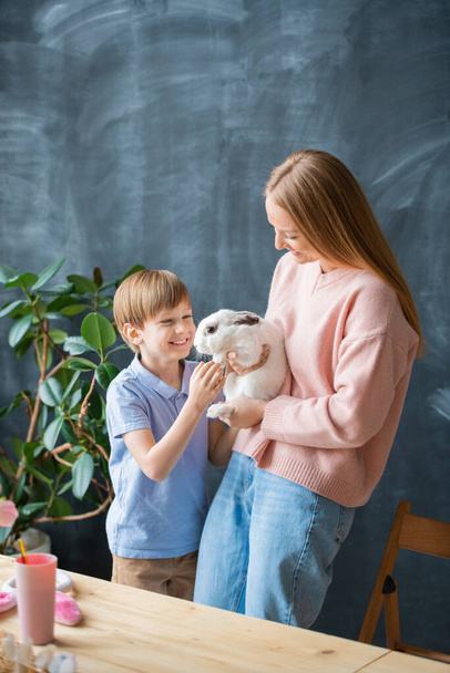 Excited cute son stroking rabbit held by mother against blackboard, Easter celebration concept - Фото, изображение