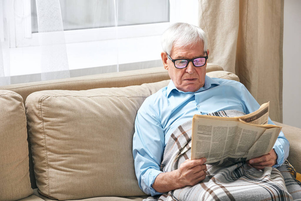 Senior grey-haired man wrapped in plaid reading newspaper while relaxing on couch by window while staying at home - Fotó, kép