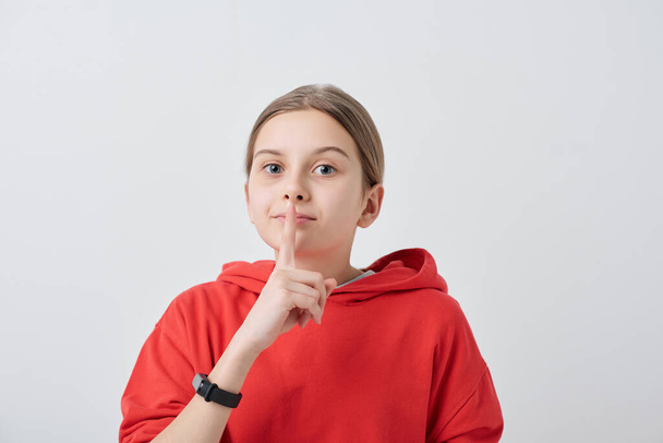 Portrait of teenage girl in red hoodie holding finger near lips while making silence sign against white background - 写真・画像