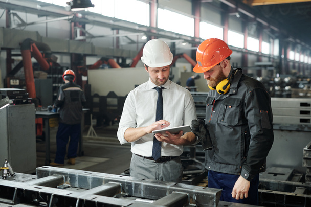 Young master in hardhat and bearded engineer discussing technical sketch on display of tablet in factory workshop - Photo, Image