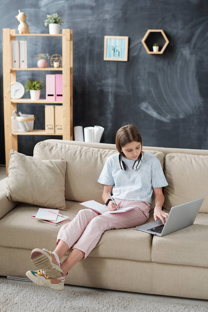 Teenage lány vezeték nélküli fejhallgató nyaka körül jegyzetel a munkafüzetben, és a laptop használata közben készül az iskolai teszt otthon - Fotó, kép