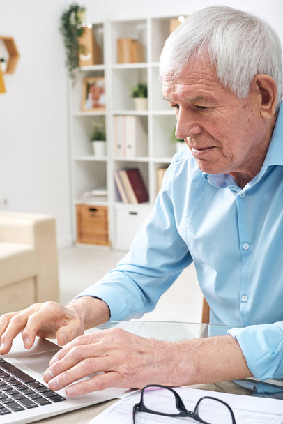 Aged retired man in blue shirt pressing keys of laptop keypad while surfing in the net for online data in home environment - 写真・画像