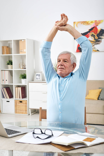Senior lekko zmęczony mężczyzna podnosi ręce nad głową podczas ćwiczeń przy stole przed laptopem podczas pracy w domu - Zdjęcie, obraz