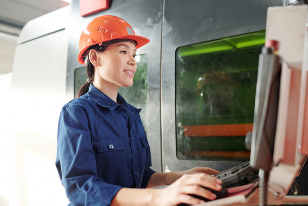 Молодая женщина-инженер в шлеме и рабочей одежде щелкает мышью, глядя на экран монитора или панели управления
 - Фото, изображение