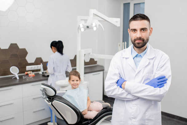 Ammattimainen hammaslääkäri valkotakissa seisoo työpaikalla hammaslääketieteellisten laitteiden taustalla, hänen avustajansa ja pieni potilas
 - Valokuva, kuva