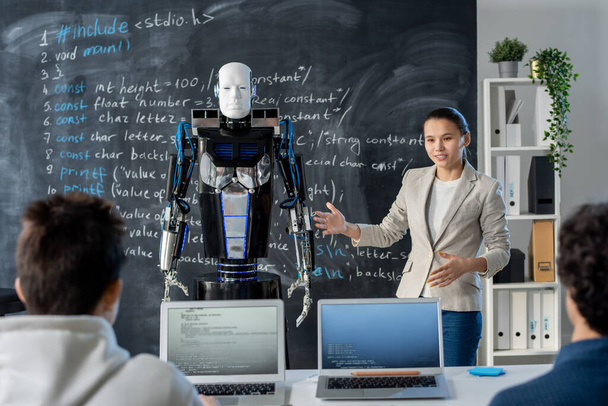 Clever young female student showing her classmates new model of automation robot while standing by blackboard - Photo, Image