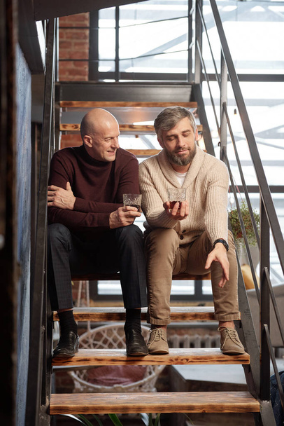 Handsome bearded man with whisky glass sitting on stairs and sharing life story with friend - Photo, Image