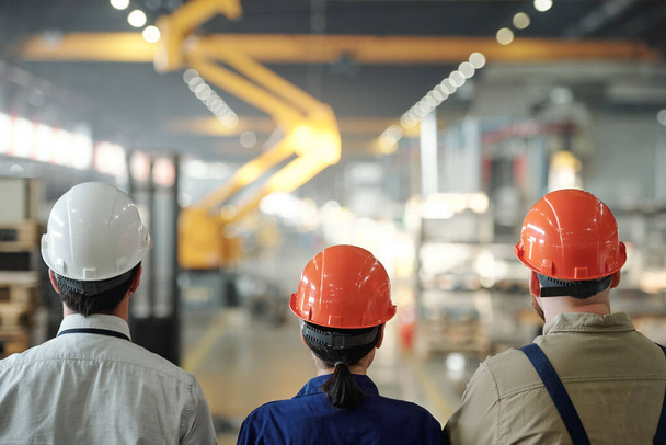 Vista posterior del equipo de ingenieros contemporáneos en cascos duros que se mueven a lo largo de una gran planta industrial frente a la cámara
 - Foto, imagen