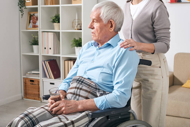 Starý zdravotně postižený muž se sklenicí vody sedí na invalidním vozíku s mladou ošetřovatelkou stojí za ním a utěšuje ho - Fotografie, Obrázek