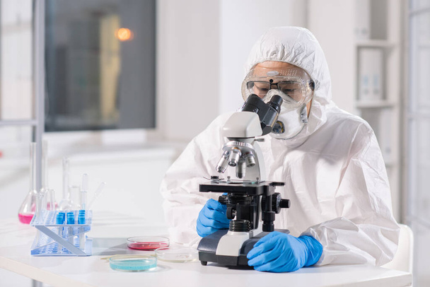 Científico de investigación de virología asiática concentrado en traje protector, máscara y respirador usando microscopio mientras analiza muestras en laboratorio
 - Foto, Imagen