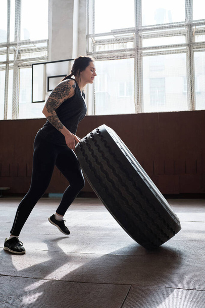 Cross-Training. Tiefansicht einer jungen, sportlich tätowierten Frau in schwarzer Aktivkleidung, die durch Reifenwechsel im Fitnessstudio Muskeln aufbaut - Foto, Bild