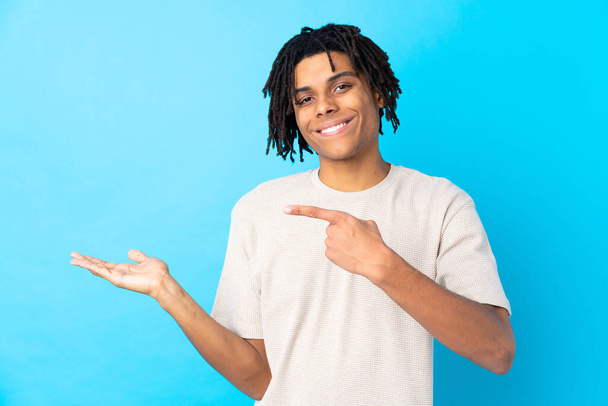 Young African American man over isolated blue background holding copyspace imaginary on the palm to insert an ad - Zdjęcie, obraz