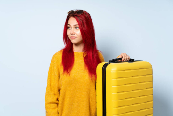 Traveler girl holding a suitcase isolated on blue background making doubts gesture looking side - Photo, image