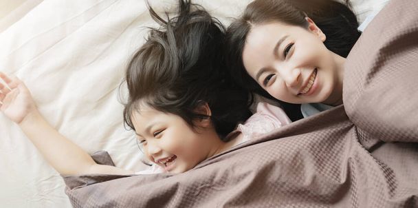 felicidade asiático família momento mãe e filha jogar e laydown com cobertor no branco cama casa interior fundo
 - Foto, Imagem