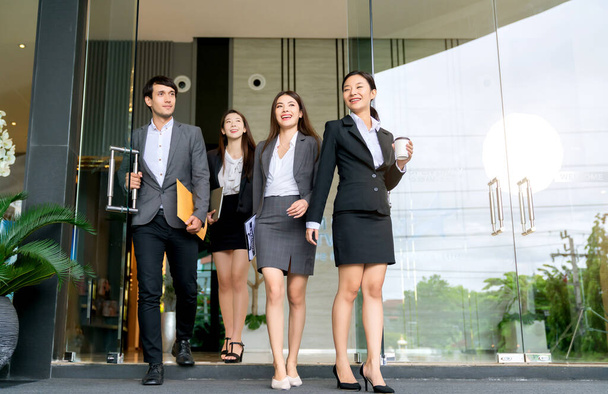 Gruppe von smarten jungen asiatischen Geschäftsleuten männliche und weibliche formelle Kleidung zu Fuß passieren moderne Büro Eingang mit Selbstvertrauen und Glück - Foto, Bild
