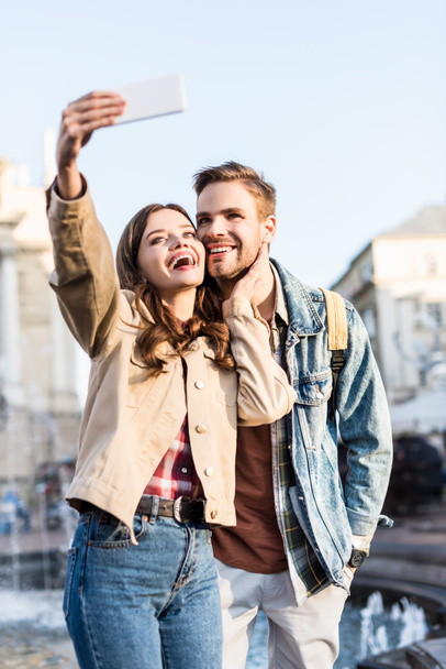 Selective focus of excited and happy couple taking selfie near fountain in city - Photo, Image