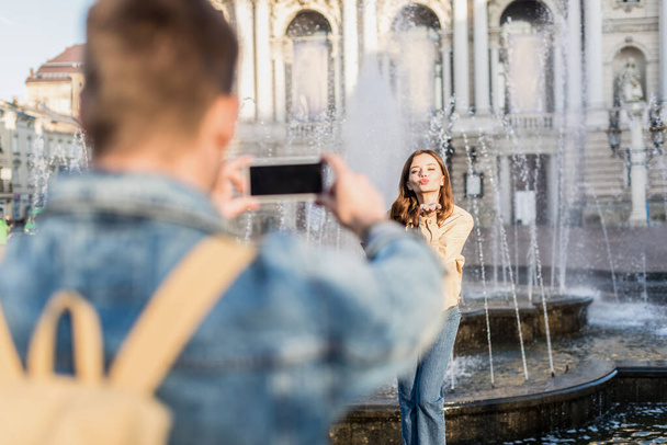 Selective focus of man taking picture with smartphone of woman blowing kiss near fountain in city - Фото, изображение