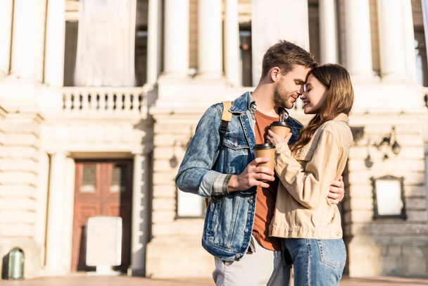 Boyfriend and girlfriend with disposable cups of coffee hugging in city - Фото, изображение