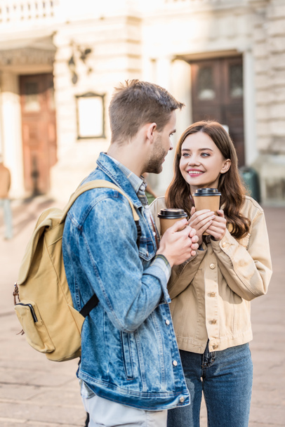 Boyfriend and girlfriend holding paper cups of coffee with clenched hands, looking at each other and smiling in city - Foto, Bild