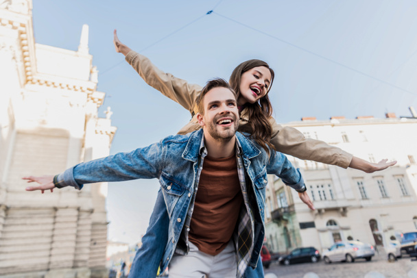 Low angle view of man smiling and piggybacking excited girl with open arms in city - Photo, Image