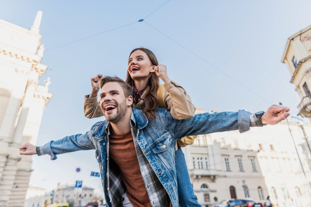 Low angle view of boyfriend smiling and piggybacking excited girl with open arms in city - Foto, Imagen