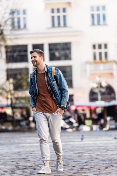 Man with backpack and hands in pockets smiling and walking in city - Foto, Bild