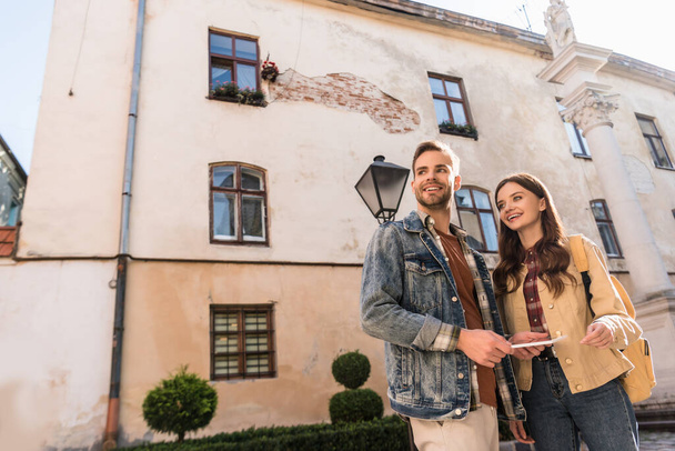 Low angle view of couple with digital tablet looking away and smiling in city - Valokuva, kuva