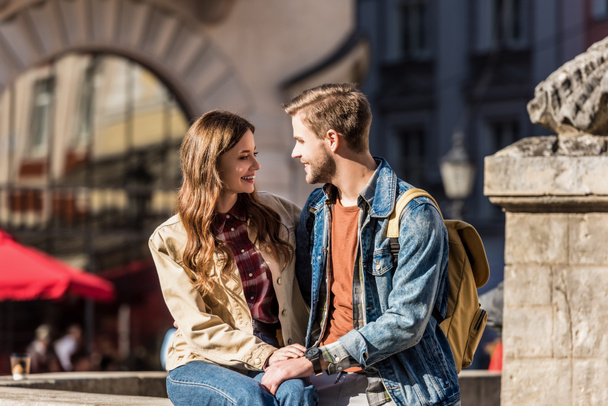 Szczęśliwa dziewczyna i chłopak siedzą razem, patrząc na siebie i uśmiechając się w mieście - Zdjęcie, obraz