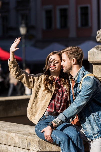 Happy girlfriend covering face with hand and boyfriend sitting together on stone and smiling in city - Foto, imagen